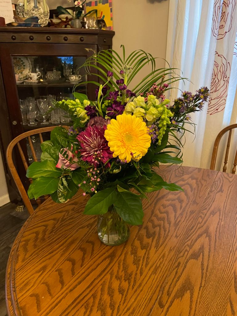 A bouquet of flowers that includes a yellow gerber daisy, magenta dahlia, purple rose, yellow reaching flowers, purple asters, and lots of greenery on a wooden table with the dining room in the background.