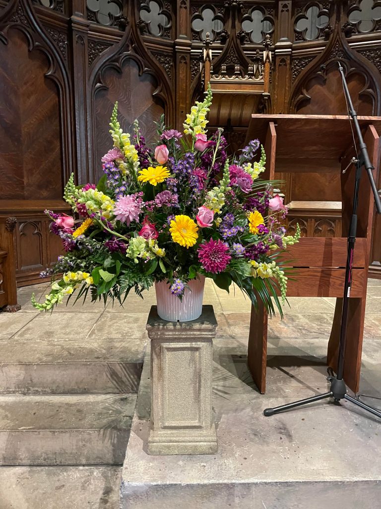 Large bouquet of flowers on a podium. Colors of flowers are yellow, pink, purple. The background has the wooden raredos of the Unitarian Universalist Church of Buffalo. The podium is to the right.