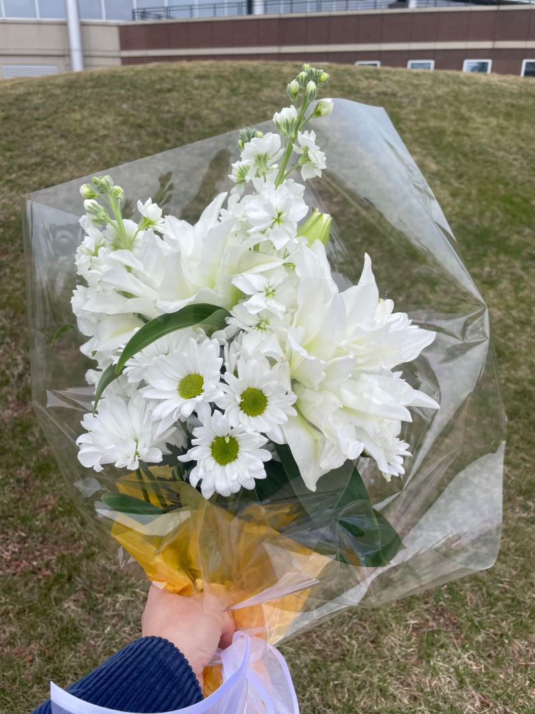 A bouquet of white flowers including white mums, white lilies, and small white flowers on long stems. They are wrapped in yellow tissue paper and clear cellophane. A lavender ribbon ties them. They are held by a left hand that had a blue corduroy jacket sleeve . The background is grass and part of an office building. 